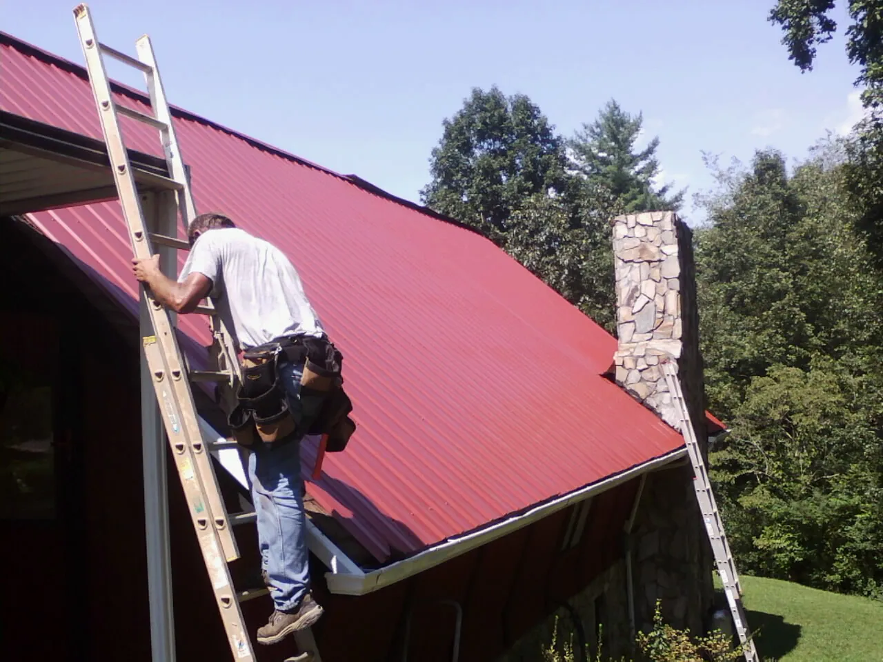 Roofer on a ladder beside a red metal roof and stone chimney — Soffit & Fascia Repair by Roofing Brothers in Purlear
